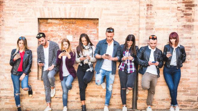 Jovens usam celular encostados em uma parede.