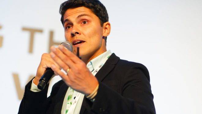 A imagem é uma foto de Eduardo Marques. Ela mostra, em plano médio, um homem que fala a um público e gesticula. O homem é branco e tem olhos escuros e cabelos curtos e escuros. Ele está vestindo um uma camisa e um blazer.