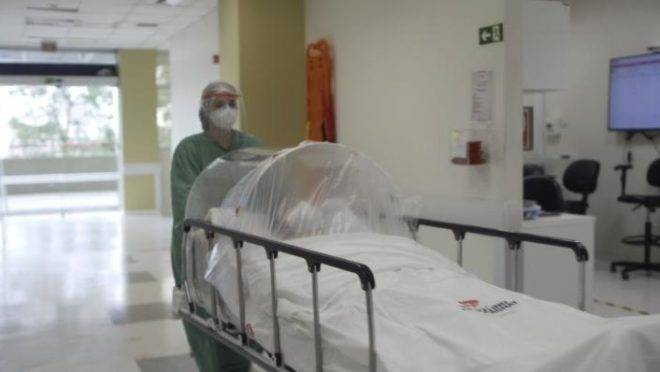 A imagem ilustra uma das ações tomadas pelo Hospital Marcelino Champagnat para lidar com a pandemia. Ela mostra uma profissional de saúde movendo um paciente Covid pelo corredor do hospital. Para não infectar outras pessoas, o paciente está em uma maca-cápsula, que o isola.