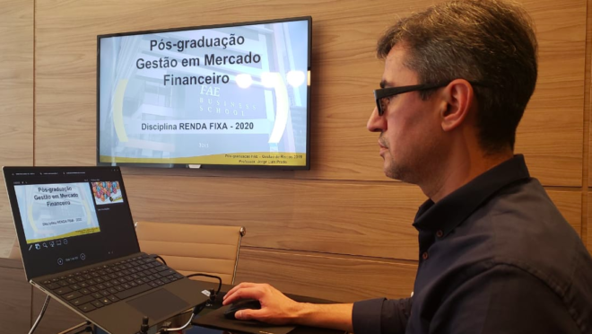A foto representa o ensino remoto emergencial adotado durante a pandemia. Ela mostra um homem, professor, de perfil, sentado em frente a um computador no qual há slides de uma aula. Ao fundo, há uma parede com uma televisão onde os slides também aparecem.
