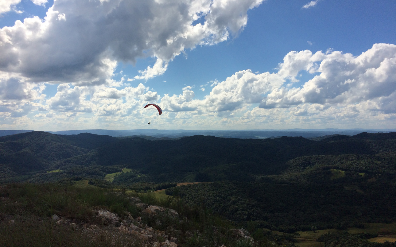 Imagem do cume do Morro do Cal. Céu azul com nuvens brancas e uma pessoa, ao longe, voando de parapente. Imagem do cume do Morro do Cal. Céu azul com nuvens brancas e uma pessoa, ao longe, voando de parapente.
