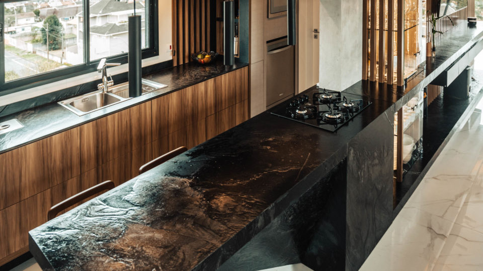 Projeto usa madeira na decoração para remeter à casa de infância do proprietário A cozinha integra o ambiente da sala de estar e jantar a partir de uma ilha. Destaca-se o uso da pedra vulcânica em preto, conversando com o estilo sóbrio e arrojado do apartamento, e as ripas de madeira.