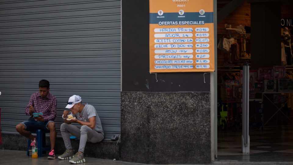Quando o governo mais atrapalha que ajuda Venezuelanos comem perto de comércio que exibe lista de preços em dólar.