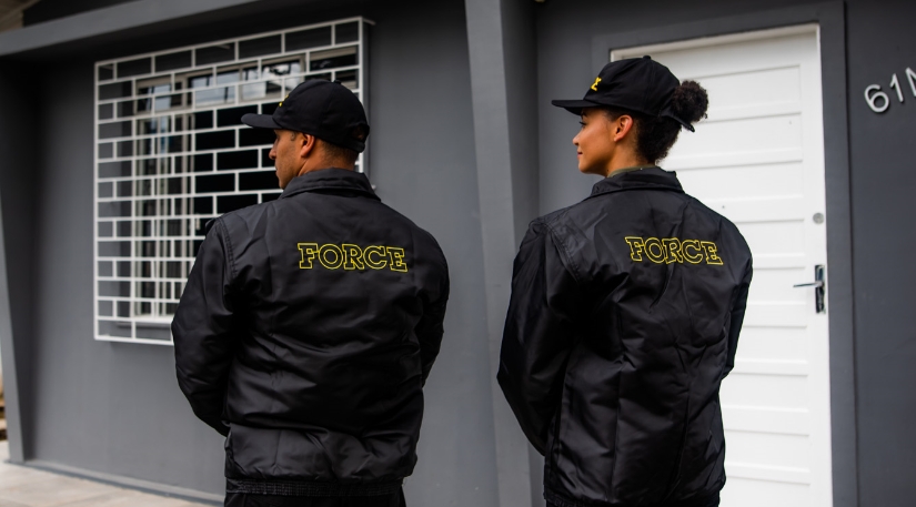 Grupo de serviços integrados compra empresa de vigilância Force Dois seguranças da empresa de vigilância Force, um homem e uma mulher, de costas, mostrando o logo da empresa na parte de trás de seus casacos.