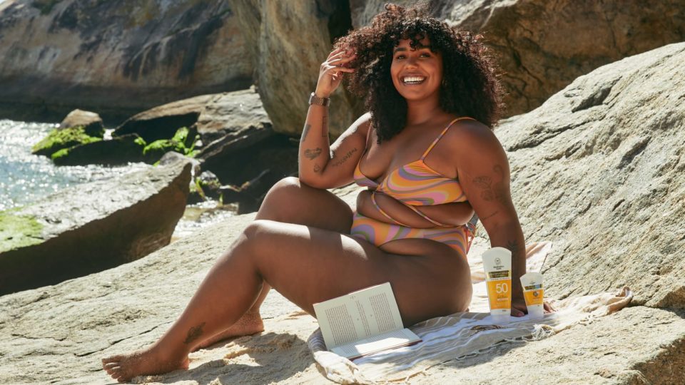 Mulher sentada na praia com um livro e o protetor solar da marca Australian Gold, do Grupo Boticário