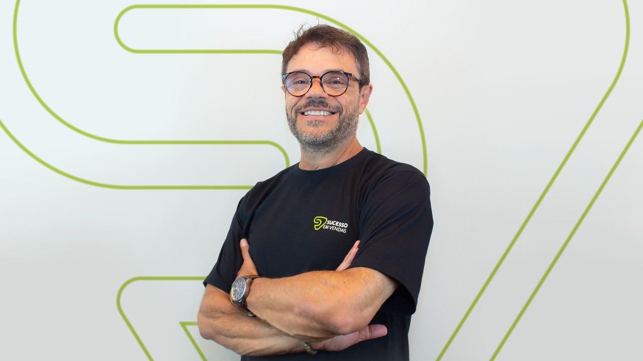 CEO da Sucesso em Vendas, Paulo Lugli, em frente a uma parede. Ele é um homem branco, com cabelos curtos, barba e óculos e aparece de braços cruzados e com uma camiseta preta.