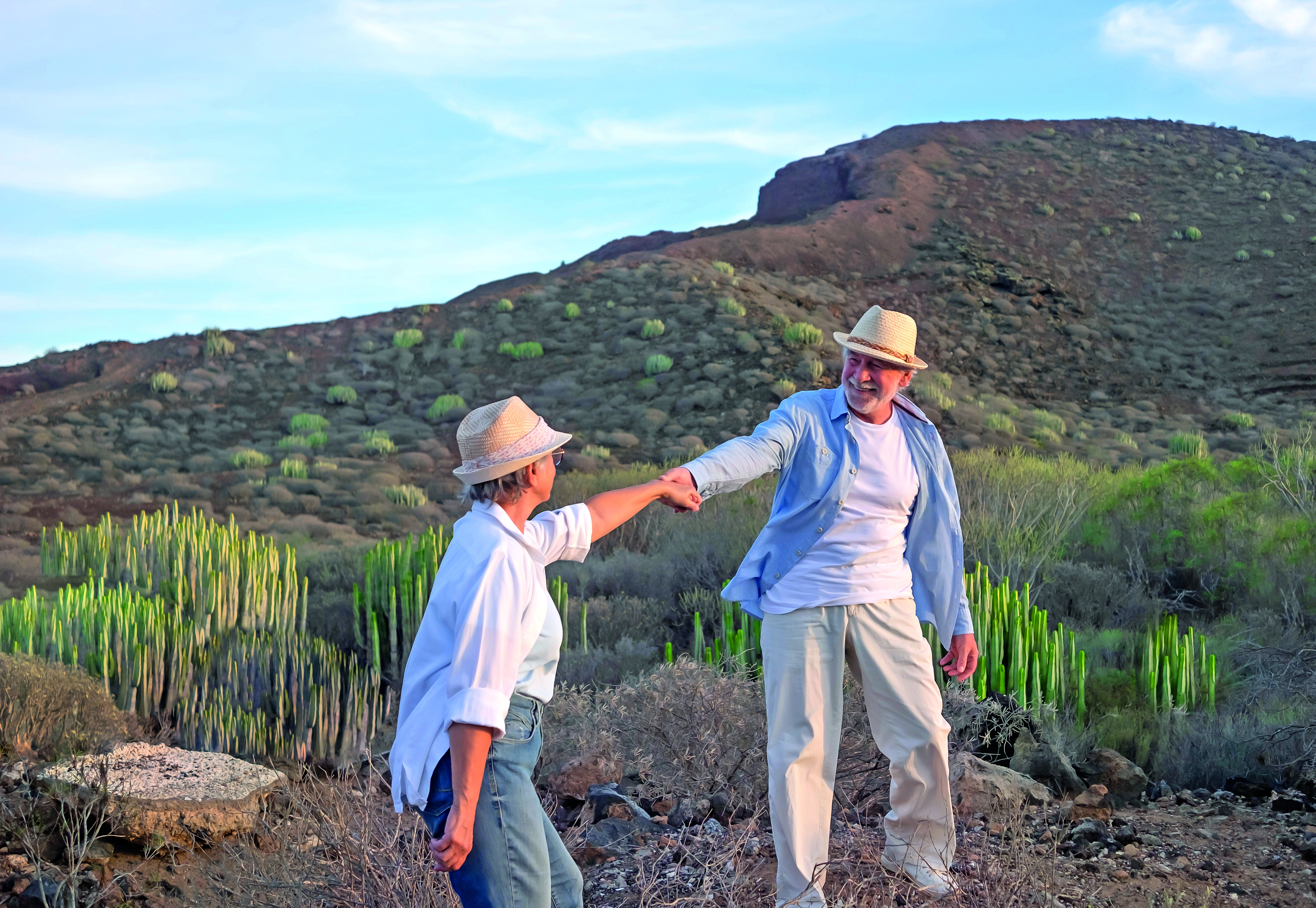 Active happy mature old senior couple of Caucasian people walking on outdoor hiking in the countryside at sunset light enjoying healthy lifestyle. The husband helping his wife to climb. Copy space