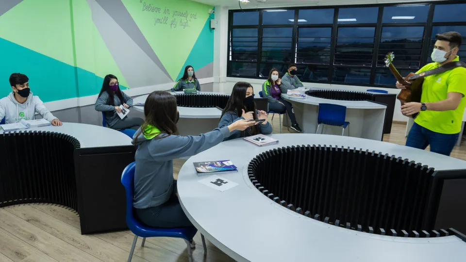 Sala de aula do Colégio Amplação, com alunos dispostos em círculos e professor tocando violão