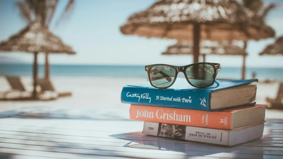 Praia. A foto mostra bangalôs que estão próximos ao mar. Em uma mesinha, estão três livros e um óculos de sol.