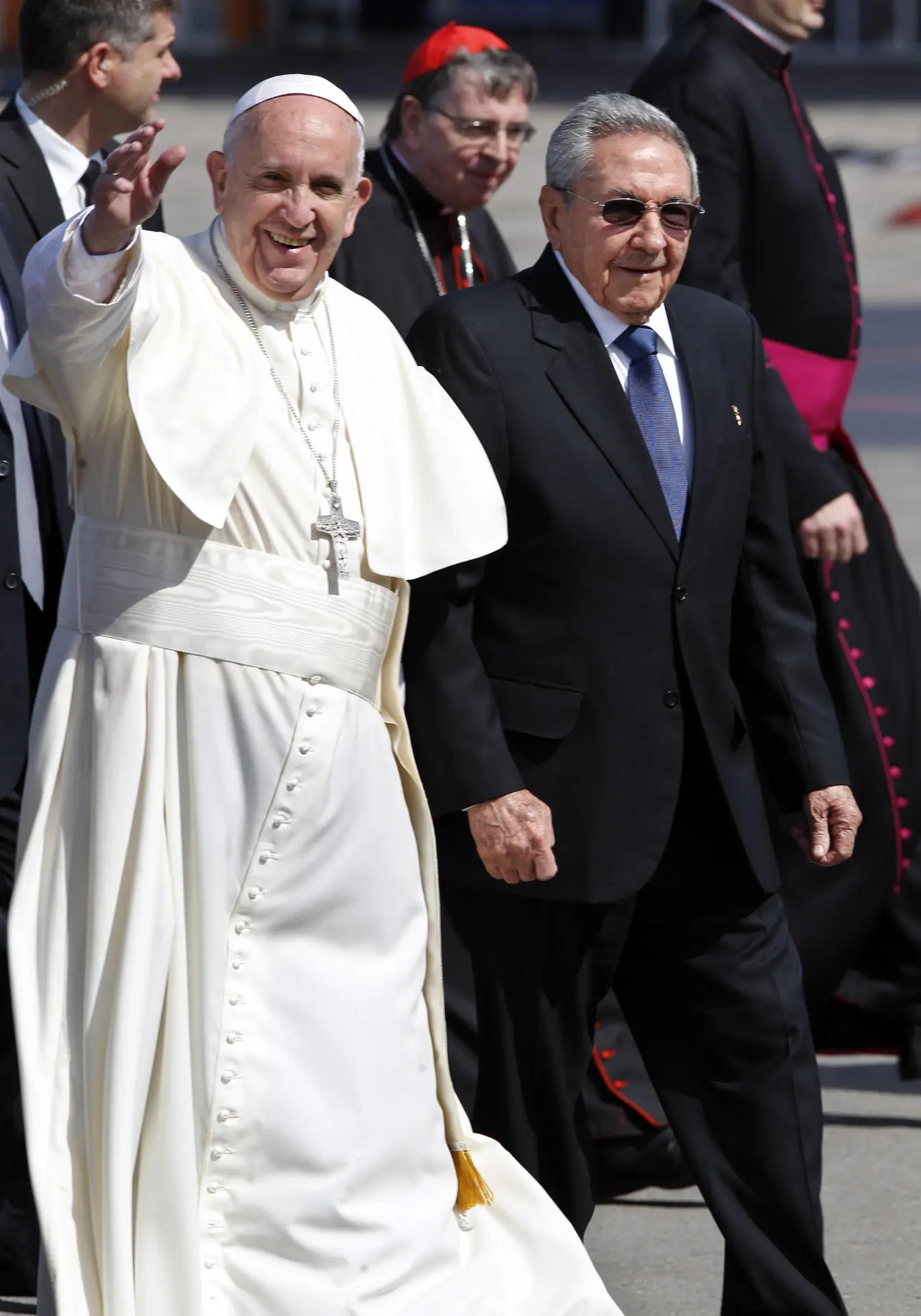 Papa Francisco ao lado de um homem vestido de terno preto com gravata azul. Ele acena para câmera e veste roupas brancas. Suas vestes deixam claro a resposta para a pergunta de quanto ganha um papa, devido sua humildade.