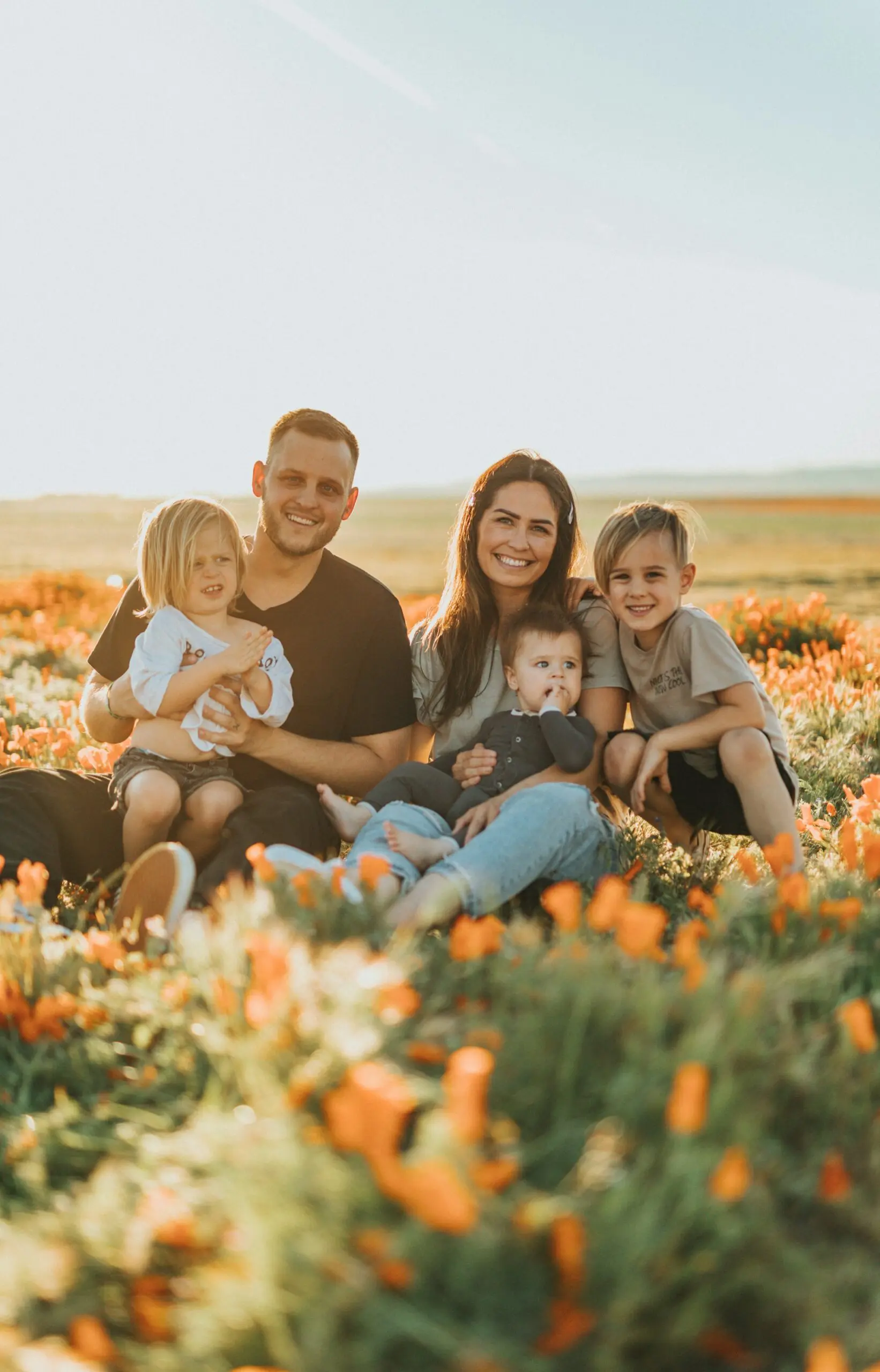 Mãe, Pai e filhos sentados em um campo de flores. Eles estão juntos e sorrindo para a foto.