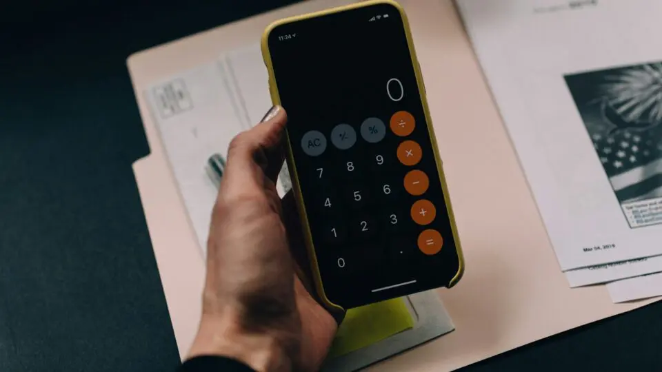 Pessoa segurando a calculadora do celular na mão. Atrás possui alguns papéis para a Declaração Anual do MEI.