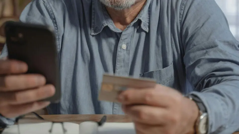 Homem segurando o celular e o cartão de crédito, pensando em suas finanças.