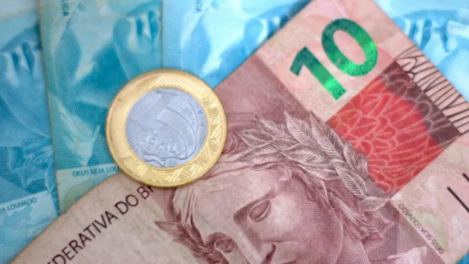 Uma nota de dez reais ao lado de várias notas de cem reais, com uma moeda de um real em cima, que fazem parte do dinheiro esquecido do Sistema de Valores a Receber.