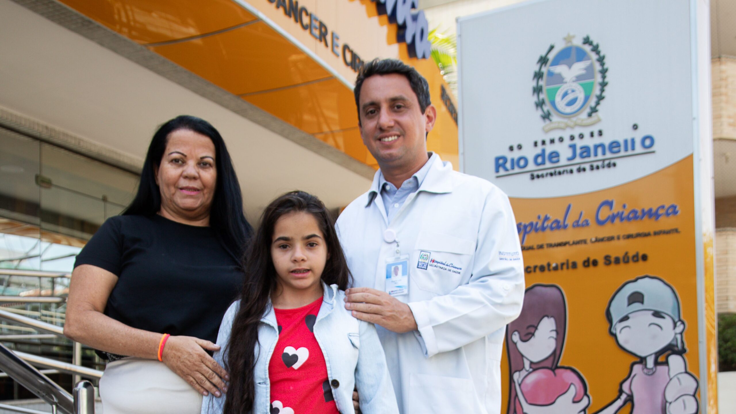 O médico Ricardo Ribas, ao lado de paciente transplantada e de sua mãe, no Hospital Estadual da Criança, símbolo da parceria com o SUS no cuidado de alta complexidade infantil.