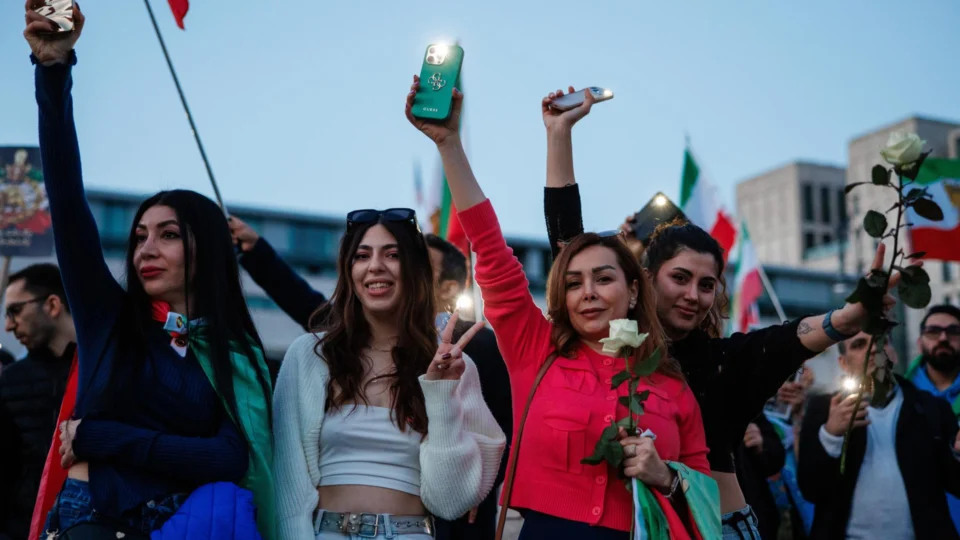 Participantes seguram flores e acendem as lanternas de seus smartphones, apontando-as para a embaixada dos EUA durante uma demonstração de apoio feita por iranianos exilados em frente à embaixada americana em Berlim, Alemanha, em 28 de fevereiro de 2026. A manifestação ocorreu sob o lema "Liberdade para o Irã"