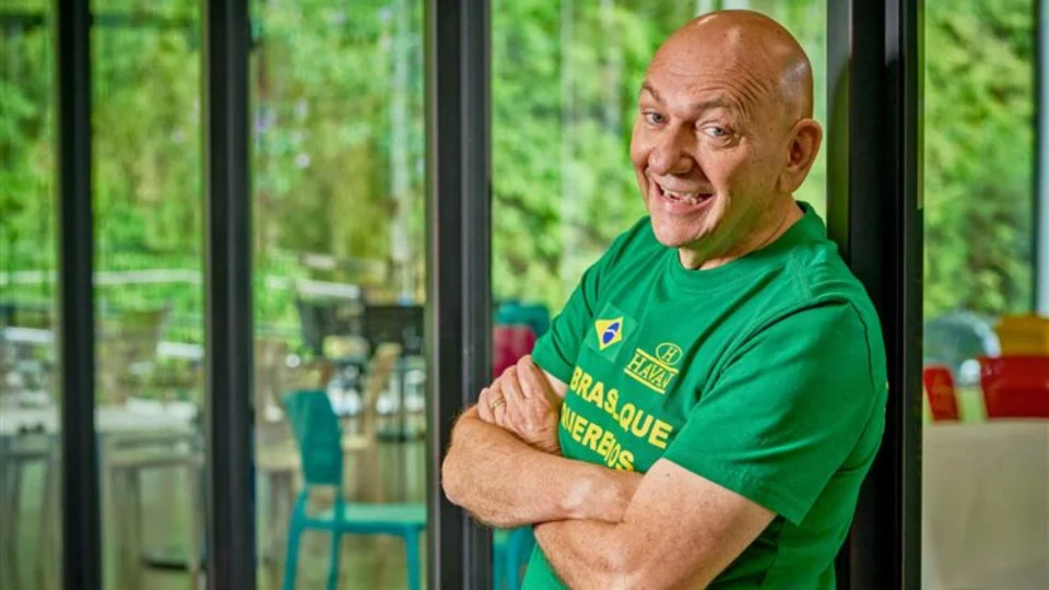 empresário dono da Havan, Luciano Hang, com a camiseta verde da loja, sorrindo, com os braços cruzados e ao fundo uma paisagem de vegetação