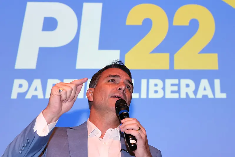 Flávio Bolsonaro com um microfone na mão apontando para frente e no fundo um painel escrito PL 22 Partido Liberal