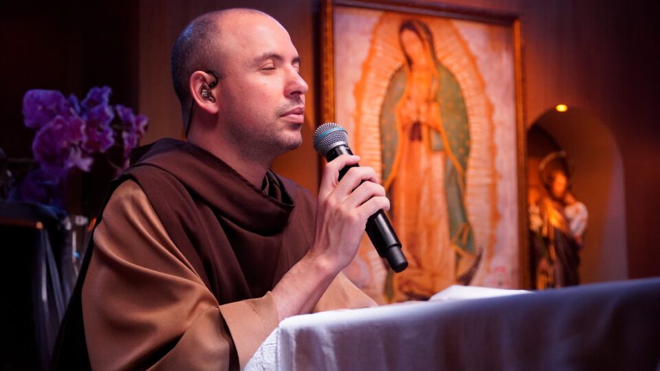 Frei Gilson ajoelhado rezando o rosário com microfone, ao fundo imagem de Nossa Senhora de Guadalupe.
