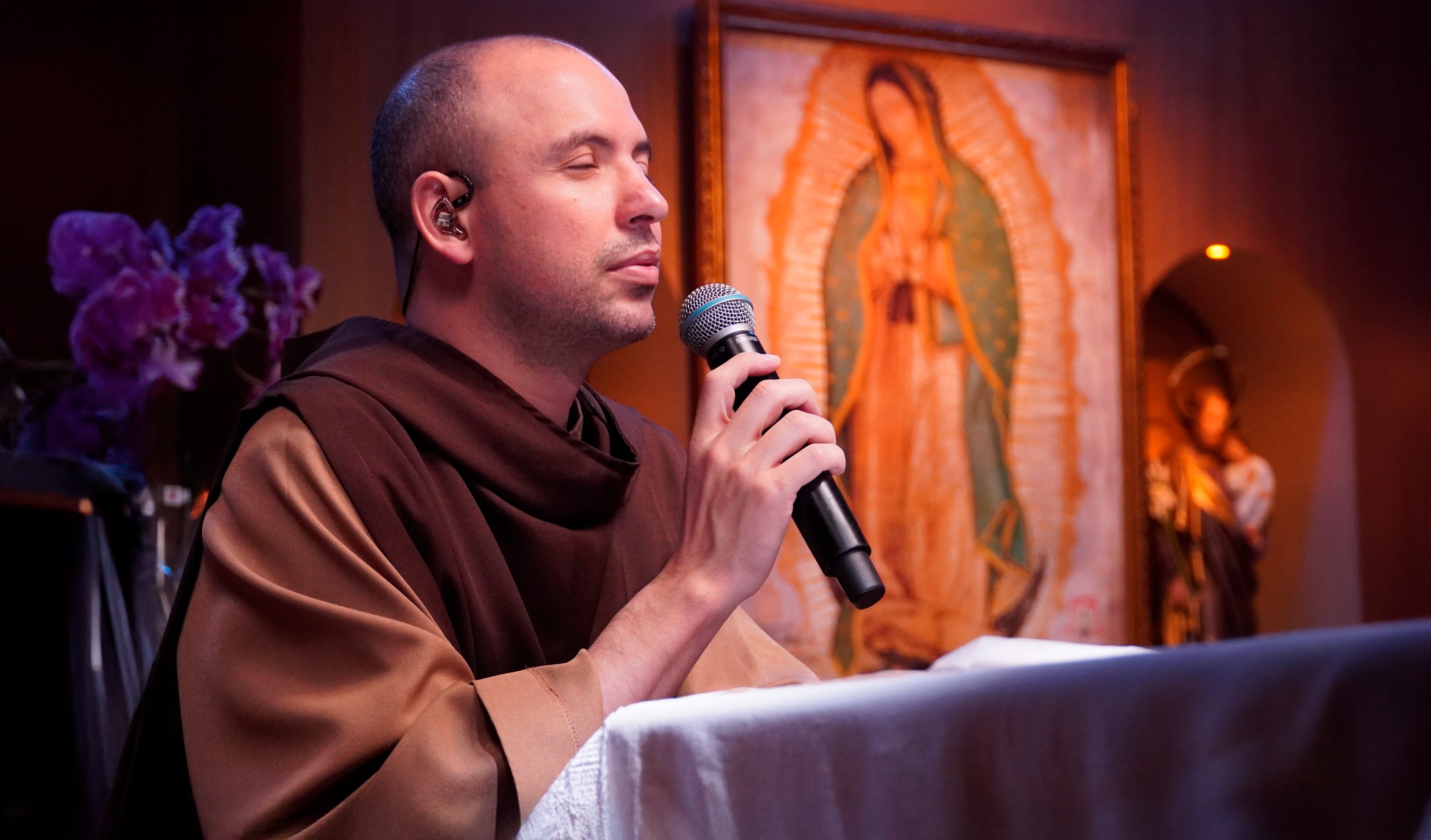 Frei Gilson ajoelhado rezando o rosário com microfone, ao fundo imagem de Nossa Senhora de Guadalupe.