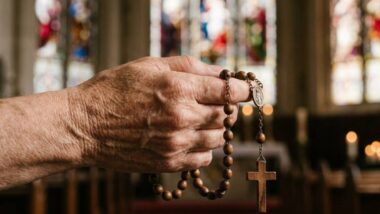 Foto em close-up de uma mão idosa e enrugada, segurando firmemente um terço de contas de madeira marrom com um crucifixo pendurado. O fundo é um interior de igreja profundamente desfocado (bokeh), mostrando a luz suave de vitrais e o brilho fraco de velas, criando uma atmosfera de devoção silenciosa.