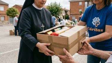 Foto documental em ambiente externo de um exchange de ajuda. Uma freira idosa em um hábito simples e véu, usando um crucifixo prateado visível, está entregando uma caixa de papelão selada de suprimentos para as mãos estendidas de um homem. Ao lado dela, uma voluntária vestindo uma camiseta azul com o logo 'Cáritas' também segura a caixa. O fundo desfocado mostra um ambiente comunitário e outras pessoas em um dia nublado.