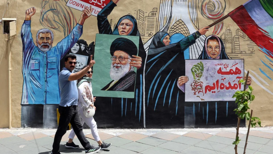 Casal iraniano caminha em frente a mural que critica ataques de Estados Unidos e Israel em Teerã, capital do país