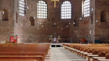 Interior de uma ampla basílica de tijolos com fileiras simétricas de bancos de madeira que levam ao altar e uma cruz dourada ao fundo