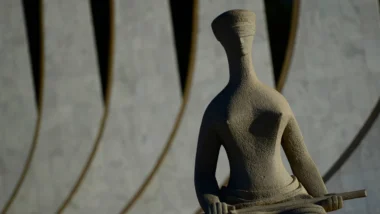Estátua da Justiça em pedra, com os olhos vendados, em close, com fundo desfocado da fachada do edifício do STF em Brasília.