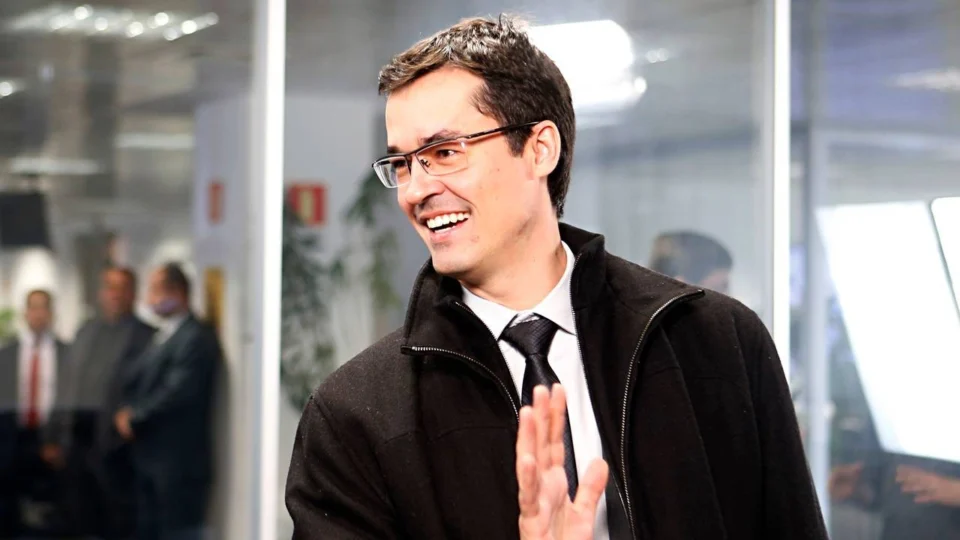 Deltan Dallagnol, candidato ao Senado pelo Paraná, sorri e acena em ambiente de escritório.