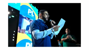 Diego Torres, de óculos e barba, fala ao microfone enquanto lê uma folha de papel em um palco à noite. Ele veste camisa polo azul-marinho sobre camisa social azul-clara. Ao fundo, um telão exibe o logotipo do PL nas cores verde, amarelo e azul. À direita, um homem de camisa verde grava a cena com o celular.