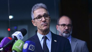 Romeu Zema, candidato do Novo à Presidência, de óculos e terno escuro, fala a jornalistas diante de microfones de emissoras, com um homem ao fundo.