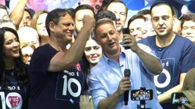 Tarcísio de Freitas ergue o punho e sorri, com camiseta azul-marinho com a inscrição "Tarcísio é 10". À esquerda dele, a primeira-dama Cristiane Freitas sorri. À direita, Guilherme Derrite, de camisa azul-clara, segura um microfone e um celular durante a ligação. Ao lado de Derrite, de camiseta azul-marinho, está o vice-governador Felício Ramuth. Ao fundo, apoiadores acompanham a comemoração.