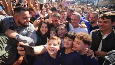 Flávio Bolsonaro e Luciano Zucco cercados por apoiadores na Expointer, em Esteio (RS), estado com seis das dez cidades de maior votação de Flávio; em primeiro plano, três adolescentes tiram uma selfie com o grupo, e ao fundo a multidão ergue celulares.
