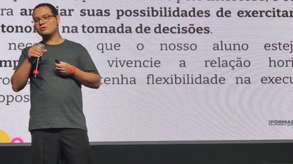 Imagem de um professor palestrando em cima de um palco, com slides no fundo.