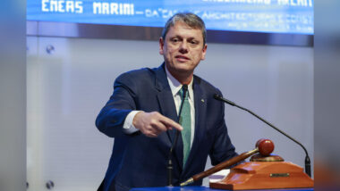 O governador Tarcísio de Freitas, de terno azul-marinho, camisa branca e gravata verde estampada, fala em um púlpito com dois microfones e aponta com a mão direita para a frente. Ao lado dele, sobre a bancada, há um martelo de madeira apoiado em uma base. Ao fundo, um painel azul com letras brancas, em leilão na B3.