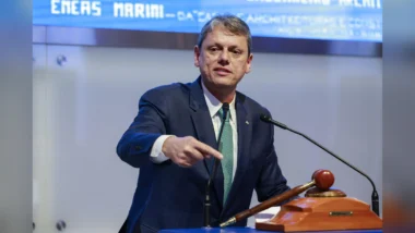 O governador Tarcísio de Freitas, de terno azul-marinho, camisa branca e gravata verde estampada, fala em um púlpito com dois microfones e aponta com a mão direita para a frente. Ao lado dele, sobre a bancada, há um martelo de madeira apoiado em uma base. Ao fundo, um painel azul com letras brancas, em leilão na B3.