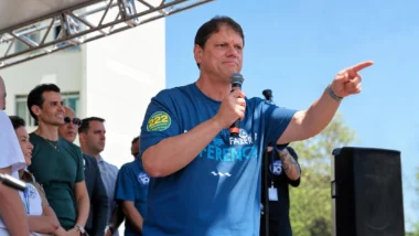 Tarcísio de Freitas fala ao microfone em um palco e aponta para o público durante evento político ao ar livre. Outras pessoas estão ao fundo, sob uma estrutura metálica, com céu azul e prédios ao redor.