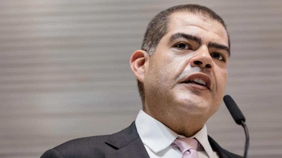 Milton Leite Filho, de cabelo escuro bem curto, fala ao microfone. Ele usa terno escuro, camisa branca e gravata rosa-clara. A foto é um close do rosto, tirada de baixo para cima, com fundo cinza listrado e desfocado.