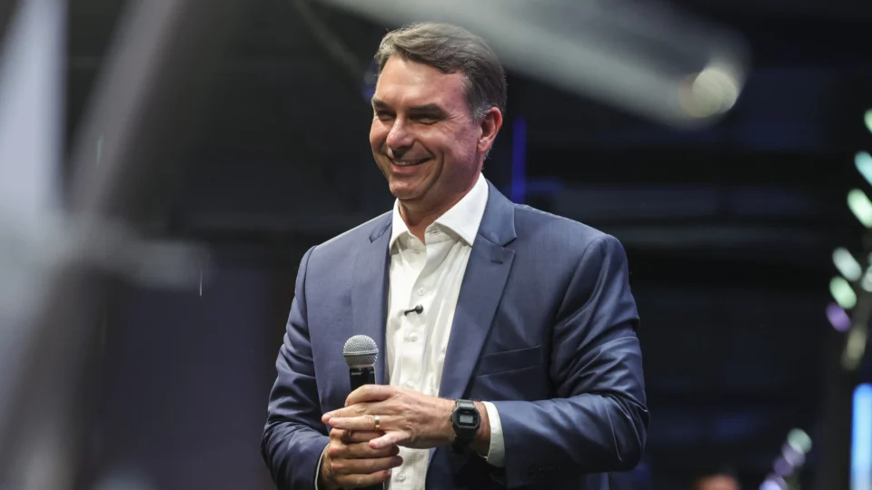 Flávio Bolsonaro sorri em pé sobre um palco, segurando um microfone com as duas mãos. Ele veste paletó azul-marinho e camisa branca sem gravata, com um microfone de lapela preso à camisa. Ao fundo, o ambiente está escuro, com luzes desfocadas em tons de verde e azul.