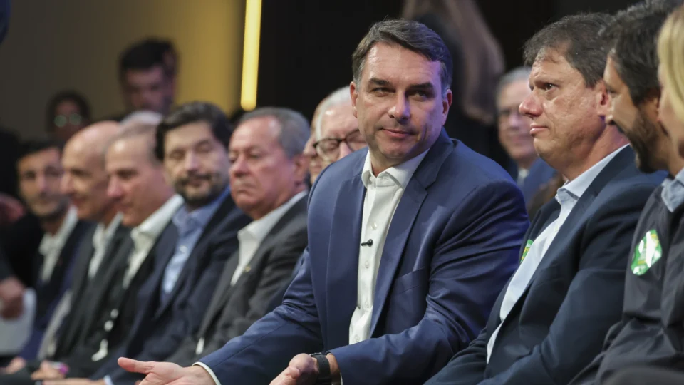 Flávio Bolsonaro, de paletó azul-marinho e camisa branca, aparece sentado em uma fileira de cadeiras, com o corpo inclinado e uma das mãos estendida, olhando para o lado. À direita dele, o governador Tarcísio de Freitas, de terno escuro e com um adesivo verde e amarelo de campanha na lapela, e, em seguida, o prefeito Ricardo Nunes, parcialmente visível. À esquerda, outros homens de terno acompanham o evento, e ao fundo há luzes desfocadas.