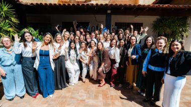Grupo de cerca de 50 mulheres posa para foto em um pátio de piso cerâmico, ao sol, em frente a uma casa de paredes bege com telhado de telhas. Muitas sorriem e acenam com a mão erguida, e várias vestem roupas brancas, azuis e pretas.