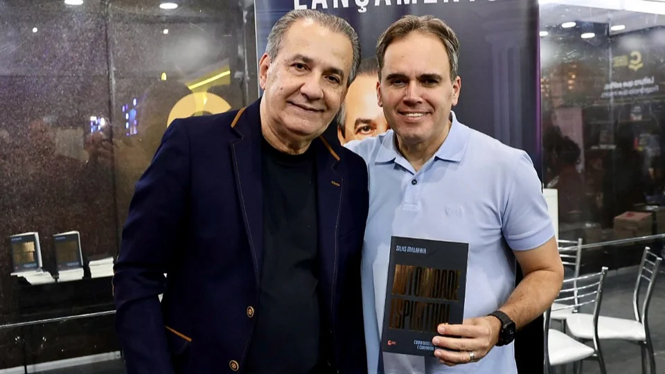 O pastor Silas Malafaia e o deputado estadual eleito Alexandre Isquierdo Malafaia posam lado a lado, sorrindo, em um estande de lançamento de livro. À esquerda, Malafaia, de cabelos grisalhos, veste blazer azul-marinho sobre camiseta preta. À direita, Isquierdo, de camisa polo azul-clara, segura o livro “Autoridade Espiritual”, de Silas Malafaia, com capa escura e letras douradas. Ao fundo, um painel com a palavra “Lançamento” e exemplares do livro expostos em uma bancada.