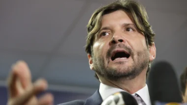 André do Prado, de cabelo castanho na altura do pescoço e barba, fala diante de microfones com a mão erguida. Ele veste terno escuro, camisa branca e gravata.