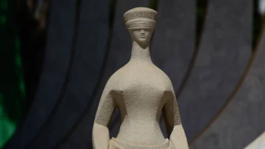 Escultura de pedra clara de uma figura feminina sentada, de olhos vendados e pescoço alongado, fotografada de perto contra um fundo escuro e desfocado com faixas curvas.