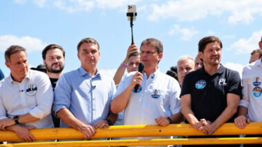 Cinco homens estão lado a lado, apoiados em uma grade amarela, ao ar livre, sob céu azul com nuvens. Tarcísio de Freitas fala ao microfone, de camisa branca. À esquerda dele estão Flávio Bolsonaro, de camisa azul-clara, e Guilherme Derrite, de camisa branca. À direita está André do Prado, de camisa polo preta com o logotipo do PL. Atrás deles aparecem outras pessoas, entre elas Filipe Sabará e o candidato a vice-presidente. Uma delas segura um celular em um bastão para filmar.
