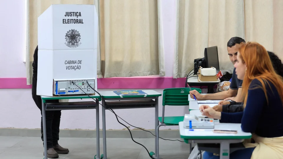 Eleitor vota em cabine com urna eletrônica enquanto mesários acompanham a seção eleitoral, em matéria sobre a abstenção no segundo turno.