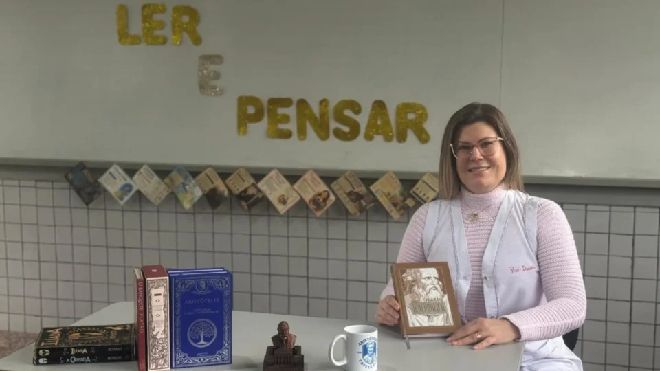 Foto da professora sentada na mesa em sala de aula com os materiais de leitura utilizados na prática.