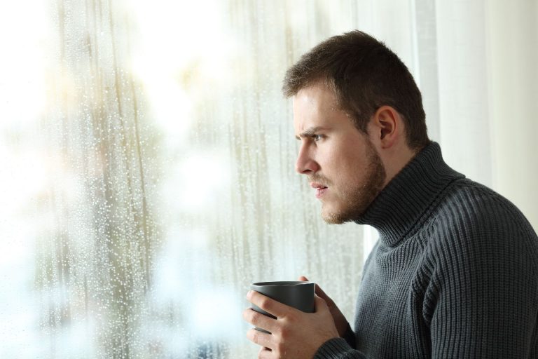 Side view portrait of an angry man looking outdoors through a window in a rainy day of winter at home